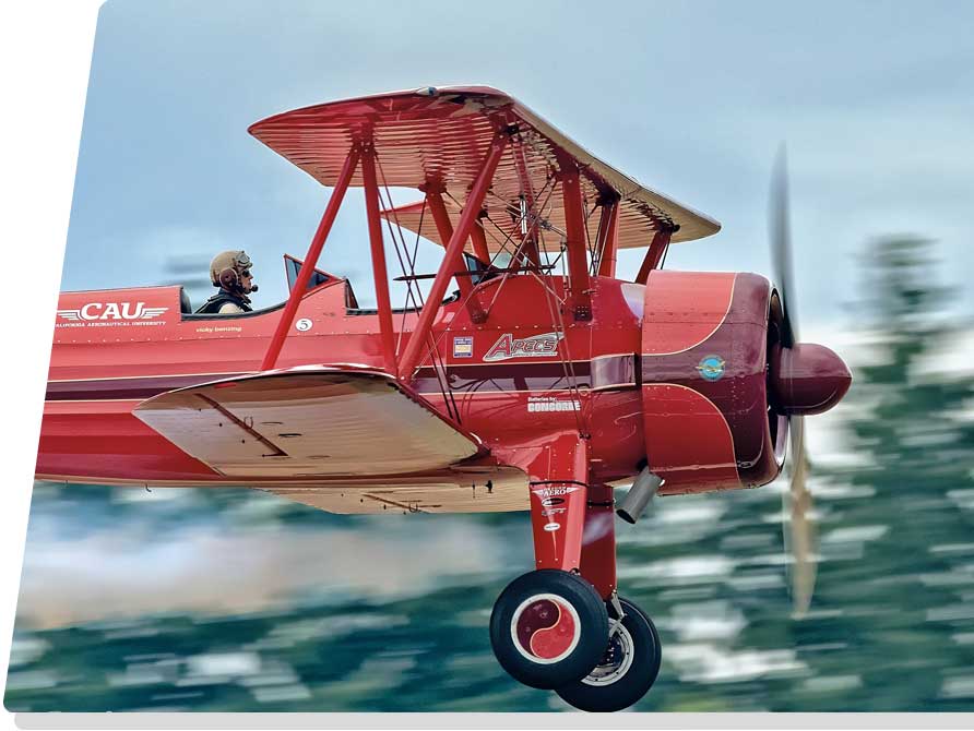 red biplane flying past pine trees